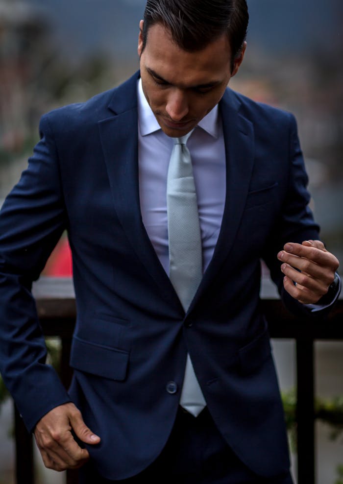 contact-img A stylish man in a dark suit looks down, showcasing elegance and sophistication, set against a blurred outdoor background.