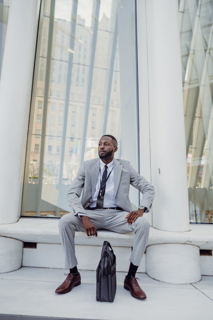about-02 Man in a suit sits outside a modern glass building, holding a laptop bag.