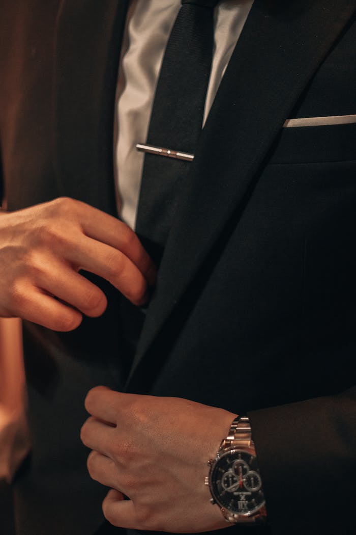 gallery-4 Close-up of a man in a suit adjusting his tie and showcasing a stylish wristwatch, capturing sophistication and style.