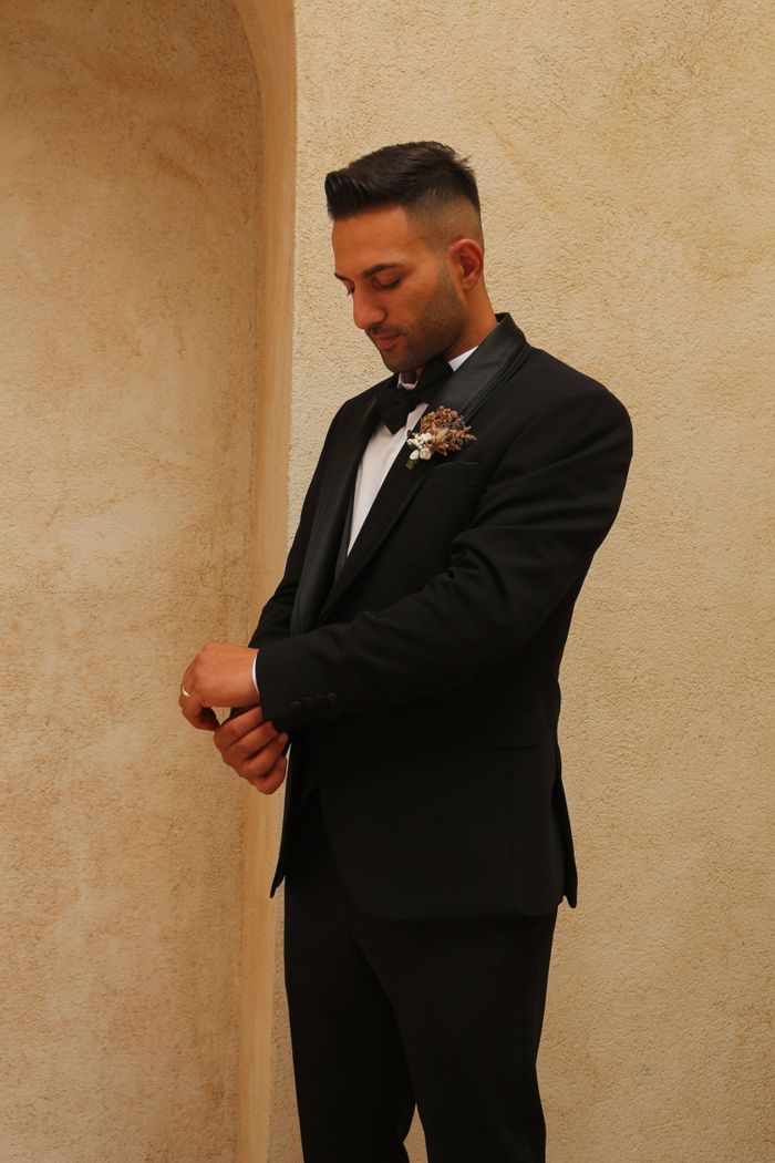 hero-img-02 A groom in a stylish tuxedo adjusts his cufflinks in a softly lit hallway.