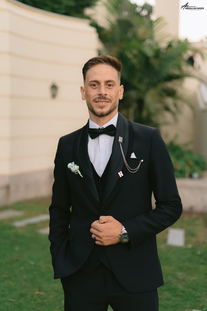 why-choose-us Man in black tuxedo with boutonniere posing confidently outside on a sunny day.