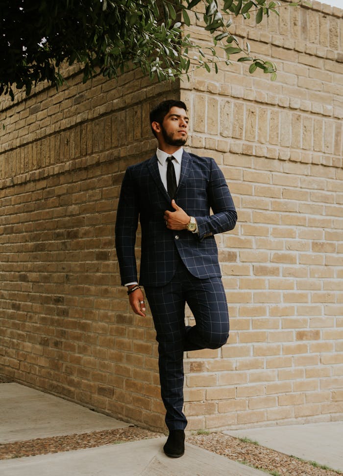gallery-2 Full body of contemplative ethnic male in stylish outfit leaning with leg on old shabby brick wall under tree on city street and looking away